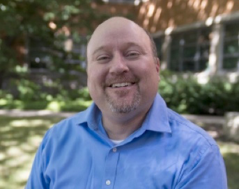 A person wearing a light blue collared shirt is seated outdoors, with green foliage and a building with large windows in the background.